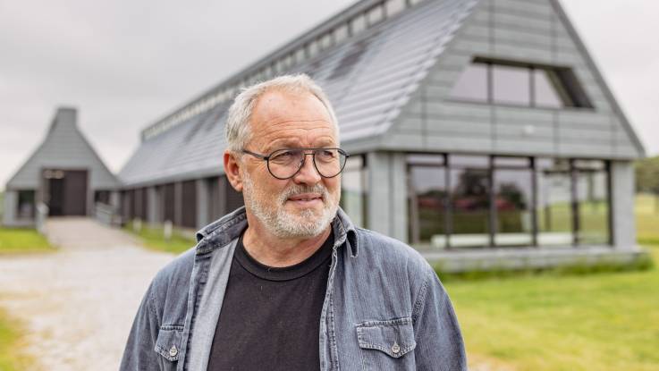 The man with glasses and gray hair gazes into the distance, lost in thought - behind him is a modern wooden house.