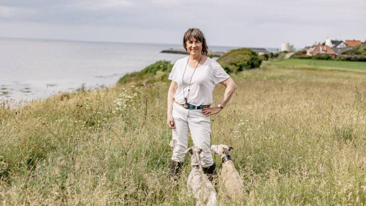A woman in white clothes and with shoulder-length hair stands on the shore, with two greyhounds sitting at her feet.