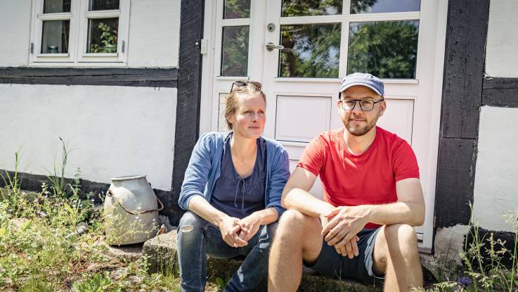 A young woman and a young man are sitting on a doorstep belonging to a white plastered half-timbered house.