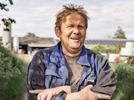 A man with a round face, blond hair and strong hands is standing outside with his arms folded and laughing.