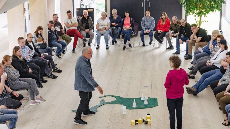 A group sits in a semicircle looking at a model of an island on which loose windmills are placed. 