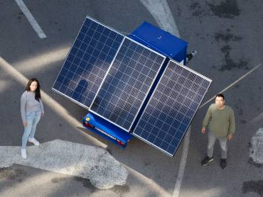 A young man and a young woman stand in front of a solar module mounted on a trailer and look friendly into the camera.