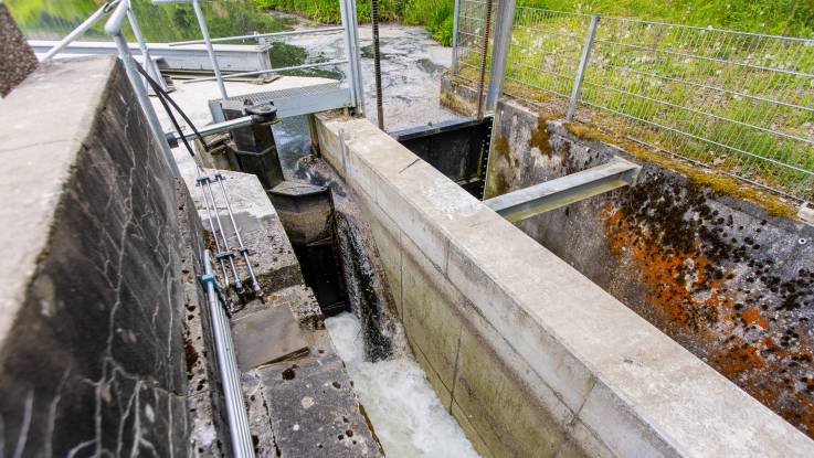 Eine kleine Schleuse öffnet sich, dunkles Wasser fließt hindurch und bildet weiße Gischt. 