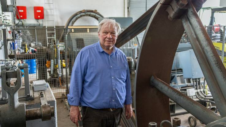 Ein älterer Mann mit grauen Haaren steht neben dem Schwungrad einer alten Wasserturbine. 