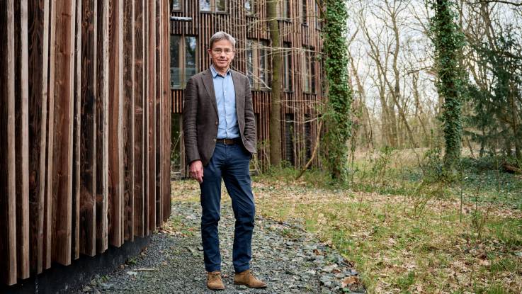 Ein Mann mittleren Alters mit grauen Haaren und randloser Brille vor einem modernen Holzhaus.
