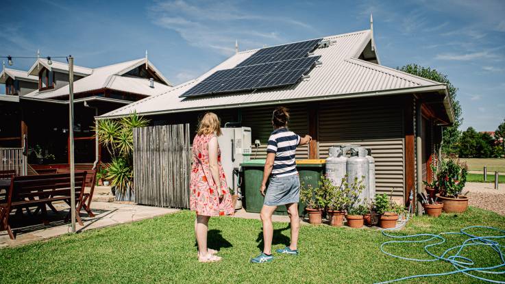 Beide Frauen stehen nun vor der Garage mit den PV-Modulen und betrachten große Gaskartuschen unterm Vordach.