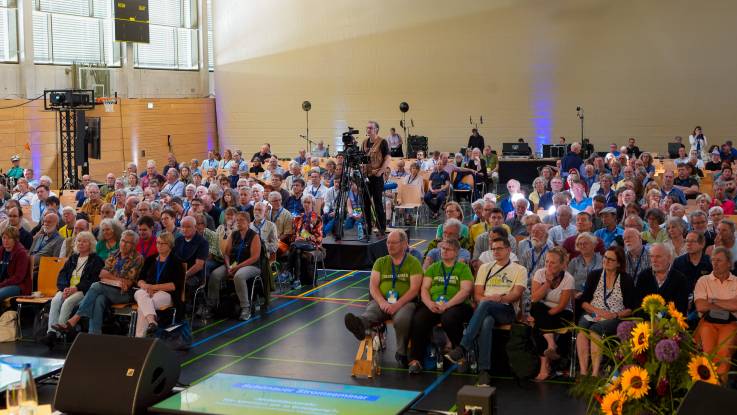 Zahlreiche Besucher sitzen aufgereiht in einer großen Sporthalle und blicken gespannt nach vorne. 