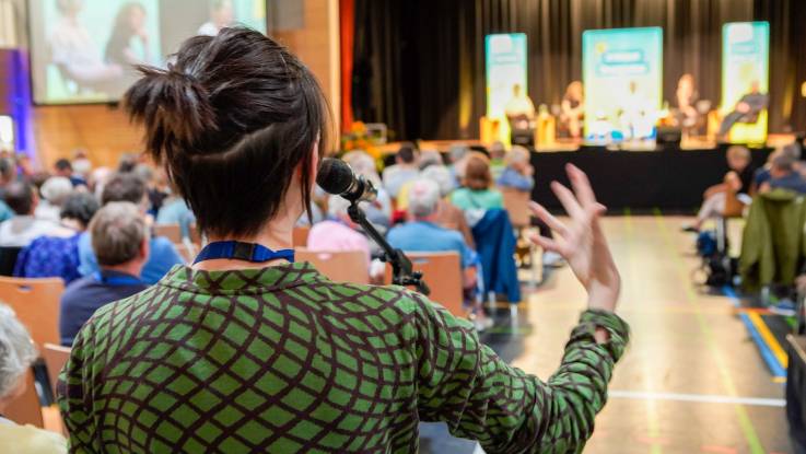 Zwischen den Stuhlreihen steht ein Standmikrophon – eine Zuschauerin stellt ihre Frage in Richtung Podium.