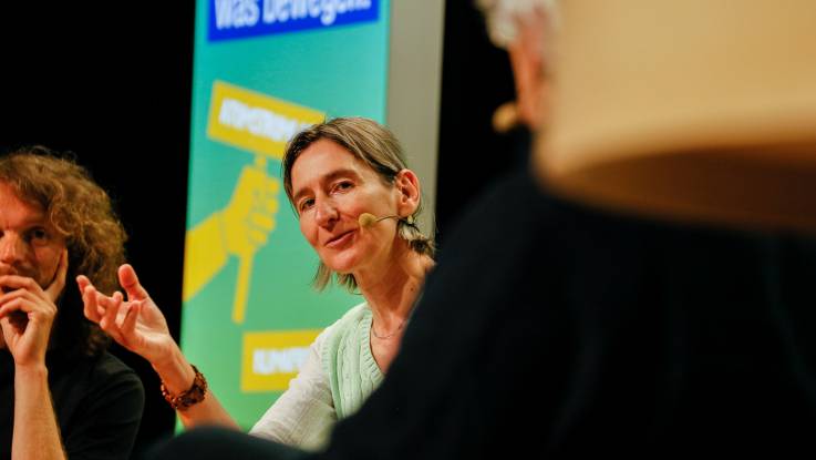 Eine Frau mit Zopf und Headset sitzt auf dem Podium während sie eine Frage an einen der Diskutanten richtet.