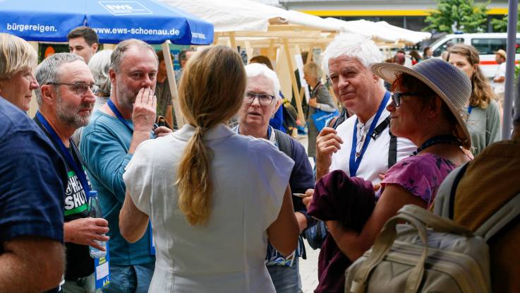 Eine blonde Frau mit Zopf steht mit dem Rücken zur Kamera, einige Besucher haben sich im Halbkreis um sie versammelt.