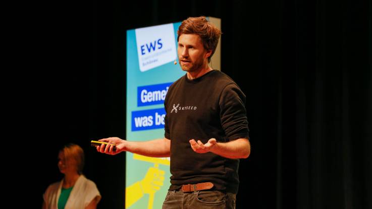 Ein Mann mit braunen Haaren steht mit ausgebreiteten Armen auf der Bühne, während er zum Publikum spricht.