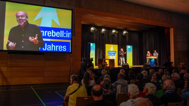 Blick auf die Bühne: Ein Mann mit Zopf hält einen Vortrag, dieser wird live auf einen großen Bildschirm übertragen.  