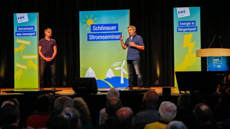 Zwei Männer stehen gemeinsam auf der Bühne, während der eine zum Publikum spricht, hört der andere aufmerksam zu.