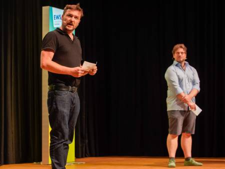 Auf der Bühne steht im Vordergrund Sebastian Sladek und spricht ins Publikum, im Hintergrund Ralf Meyer in kurzen Hosen.