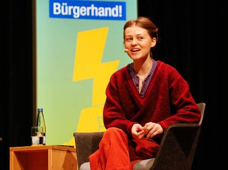 Eine junge Frau in weiter Kleidung und hochgesteckten Haaren spricht auf dem Podium.