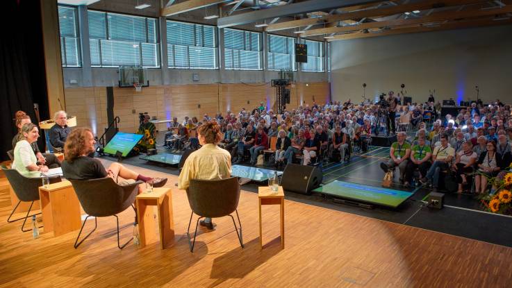 Die Podium sitzt im Halbkreis auf der Bühne, während das Publikum der Diskussion folgt.