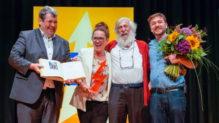 Vier lachende Personen: während die einzige Frau einen blitzartigen Preis hält, präsentiert einer der Männer ein goldenes Buch.