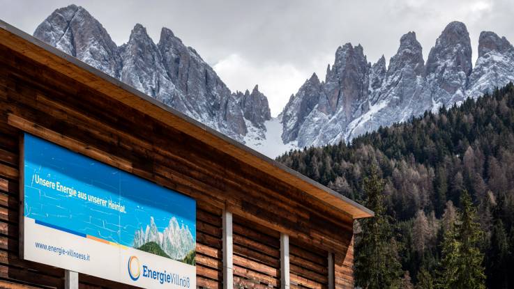 Behind a wooden house stretches a slope full of fir trees, from which a jagged chain of peaks rises. 