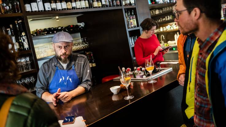 An innkeeper with a slouch hat and an apron chats with his guests while standing behind the counter. 