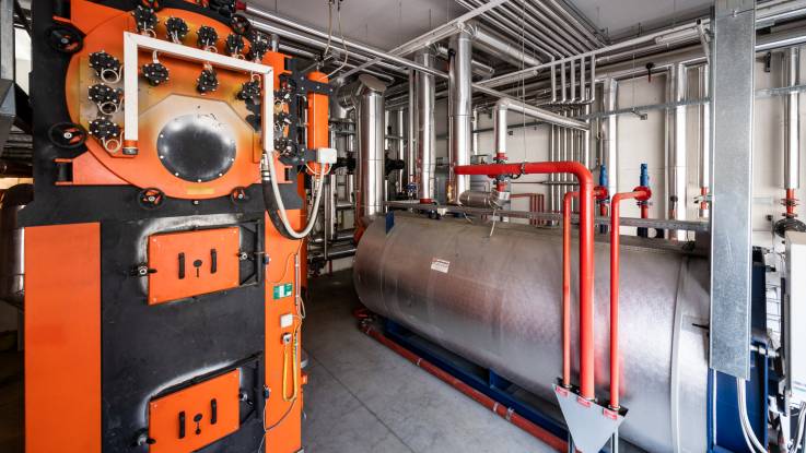 There are two boilers in a boiler room: numerous pipes run out of them and along the ceilings.