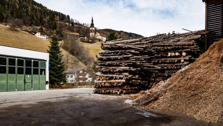 In the yard of the heating plant, tree trunks are stacked as high as a house, and wood chips lie next to them under a canopy. 