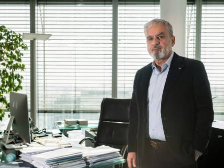 A man in a suit with grey hair and a beard is standing in the office - documents are piled up on the desk next to him. 