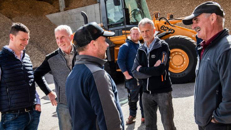  A group of men stand together, an orange excavator parked behind them in front of a pile of wood chips.