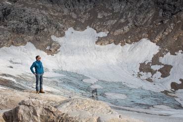 Ein Mann in blauer Jacke steht in einem Talkessel und blickt in die Ferne – unter dem Schnee kommt Geröll zum Vorschein.
