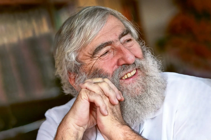 Ein betagter Mann mit grauen Haaren und Rauschebart legt seinen Kopf in die seitlich gefalteten Hände und lächelt.