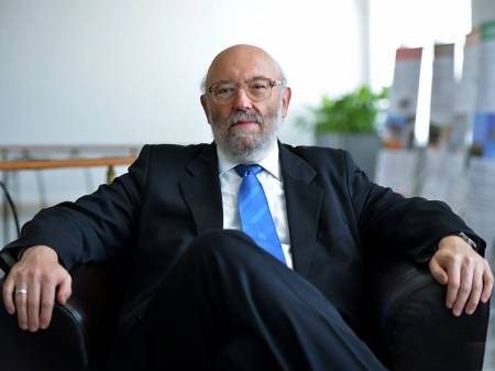 Ein älterer Mann mit Glatze und grauem Vollbart sitzt mit überschlagenen Beinen in einem Sessel.