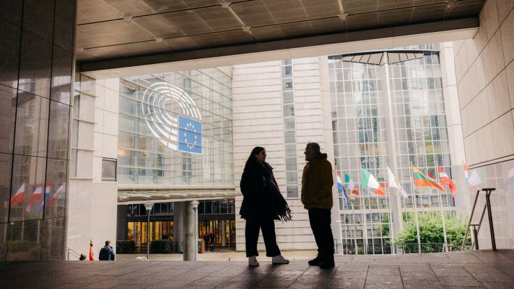 The woman and the man are now standing outside, with European flags in the courtyard behind them - one façade is emblazoned with the European flag.