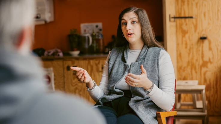 A woman in her early thirties with long brown hair and an alert gaze sits opposite a much older person. 