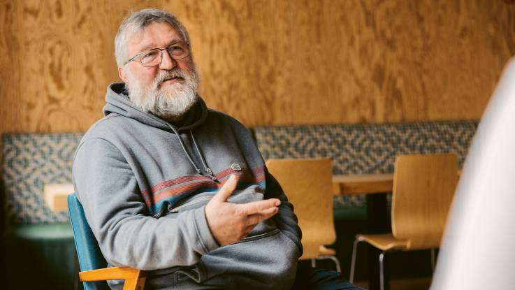 An older, friendly-looking man with gray hair and glasses sits in a hoodie on a chair. 