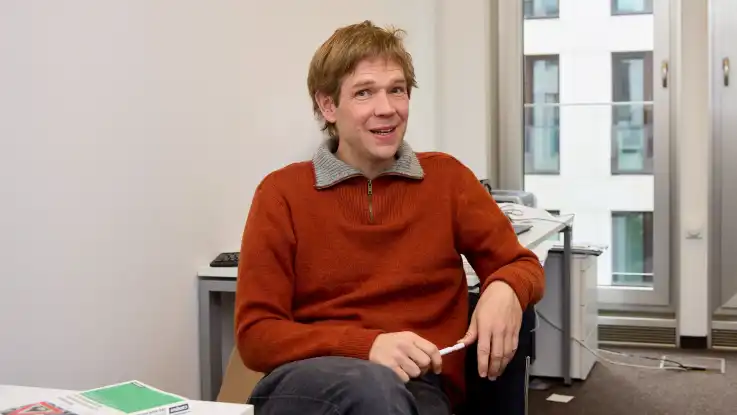 Der dunkelblonde Mann sitzt nun in einem Büro – hinter ihm steht ein Schreibtisch, vor ihm liegt ein Buch.