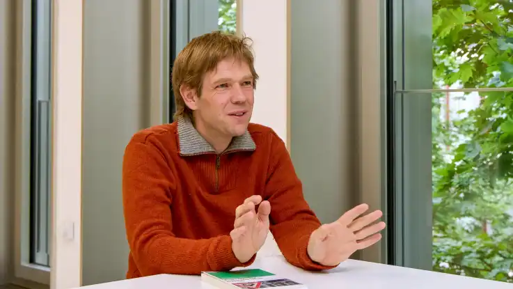 Der Mann gestikuliert, während er angeregt erzählt. Mittlerweile sitzt er an einem Fenster, durch das das satte Grün eines Baumes zu sehen ist.