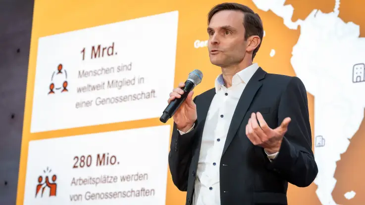 Ein Mann mit Kurzhaarschnitt steht vor einer großen Leinwand und spricht in ein Handmikrophon.