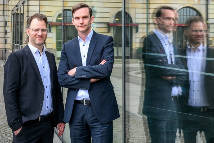 Zwei Männer mittleren Alters in schwarzen Anzügen und weißen Hemden posieren freundlich für die Kamera. 