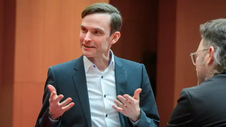 Beide Männer unterhalten sich: Der Mann mit dem grau melierten Bart ist seitlich von hinten zu sehen, während er den Ausführungen seines Gegenübers lauscht.