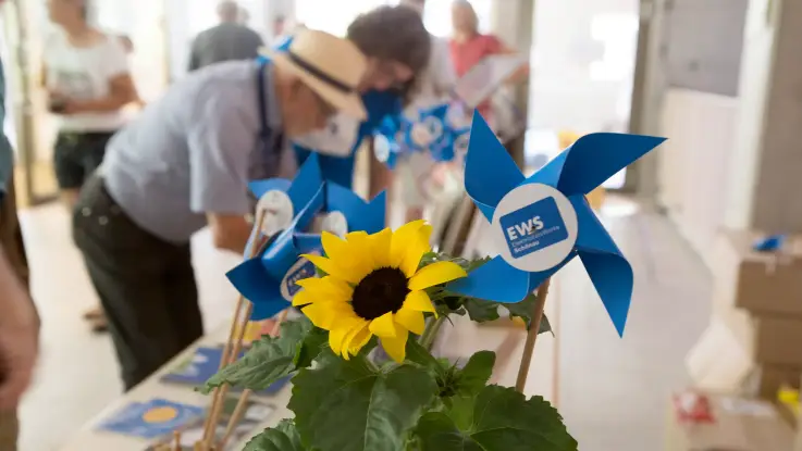 Eine Sonnenblume und kleine, blaue Spielzeugwindräder auf einem Tisch – dahinter sind unscharf Personen zu sehen. 