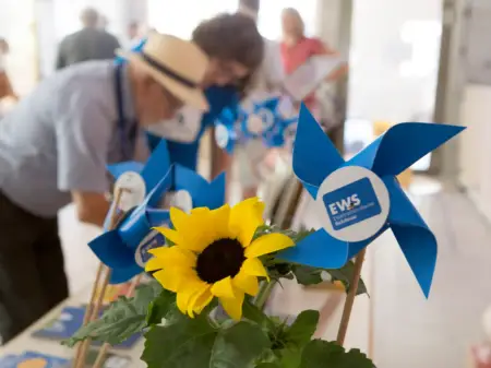 Eine Sonnenblume und kleine, blaue Spielzeugwindräder auf einem Tisch – dahinter sind unscharf Personen zu sehen. 