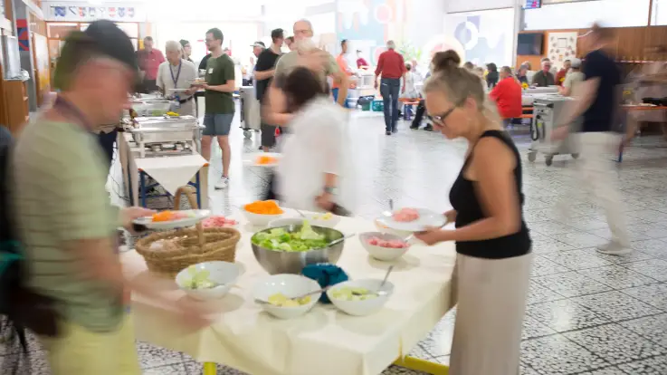 Zahlreiche Menschen bedienen sich an einem Büffet – im Hintergrund sitzen einige bereits zu Tisch und essen. 