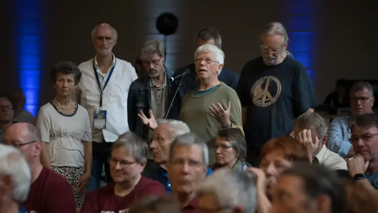 Zwischen den Zuschauerrängen haben sich einige Personen hinter einem Standmikro aufgereiht. 