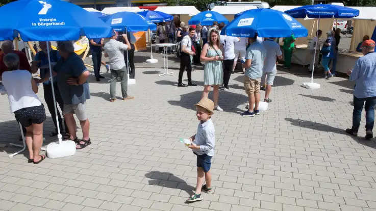 Ein kleiner Junge mit Hut steht etwas verloren zwischen Erwachsenen, die unter blauen Sonnenschirmen Schutz suchen. 