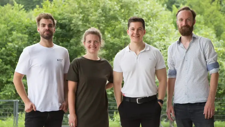 Drei Männer und eine Frau stehen nebeneinander im Freien – den Hintergrund bildet eine grüne Front aus Bäumen. 