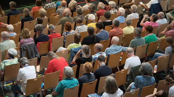 Von oben: Im Innern einer Halle sind die Sitzreihen gefüllt, einige Personen schauen erwartungsvoll nach vorne.