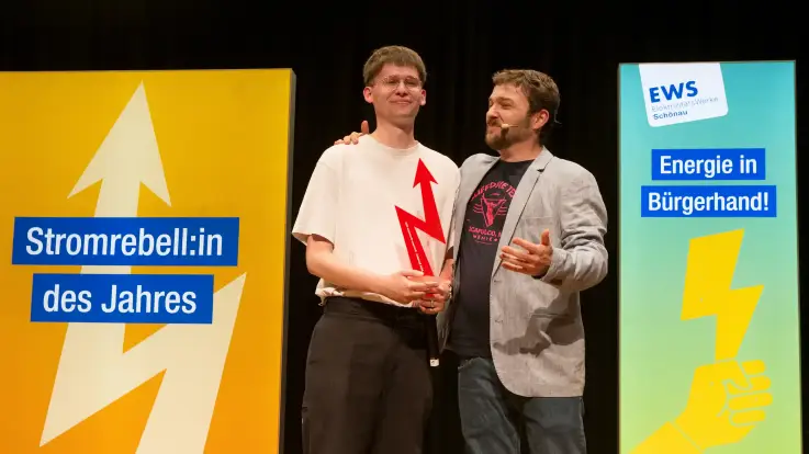 Ein junger Mann hält eine Trophäe und steht Arm in Arm mit einem etwas älteren Mann – auf dem Banner neben ihm steht: Stromrebell des Jahres. 